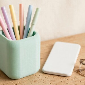 May include: A mint green square pen holder containing six pastel-coloured pens in shades of pink, purple, yellow, peach, blue, and light green. Next to the holder is a plain white rectangular smartphone and a pair of clear-framed eyeglasses.