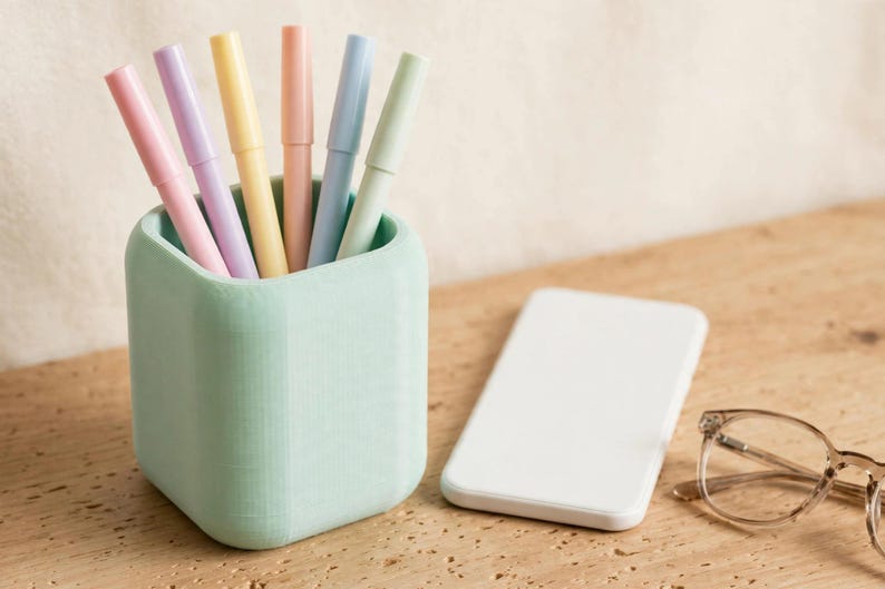 May include: A mint green square pen holder containing six pastel-coloured pens in shades of pink, purple, yellow, peach, blue, and light green. Next to the holder is a plain white rectangular smartphone and a pair of clear-framed eyeglasses.