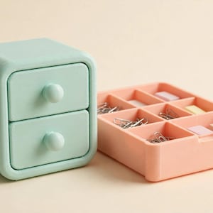 May include: A mint green two-drawer mini storage chest with rounded corners and spherical knobs, positioned next to a peach-coloured rectangular desk organiser tray featuring nine individual compartments filled with metal paper clips.