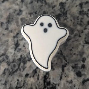 May include: A small, white, ghost-shaped charm with a black outline. The ghost features two circular black eyes and a smaller circular black mouth, creating a simple, recognisable spectral figure with a smooth, matte-like surface texture.