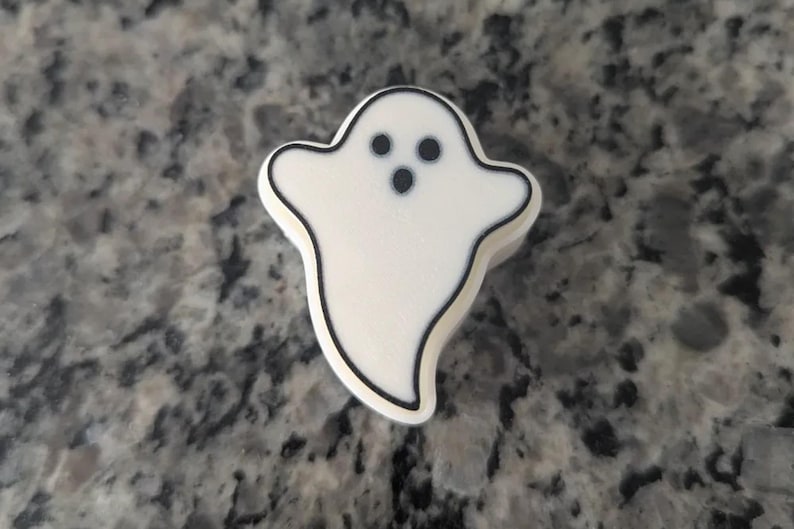 May include: A small, white, ghost-shaped charm with a black outline. The ghost features two circular black eyes and a smaller circular black mouth, creating a simple, recognisable spectral figure with a smooth, matte-like surface texture.
