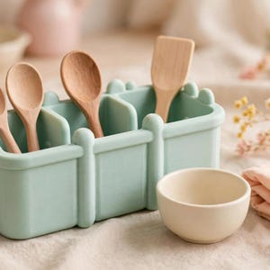 May include: A light green rectangular silicone organiser with three divided compartments containing three light brown wooden spoons and one light brown wooden spatula, accompanied by a small, round, off-white ceramic bowl.