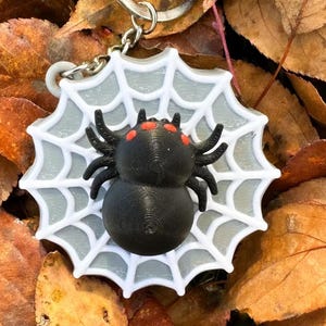 May include: A circular keyring featuring a white spider web design with a grey centre. A black spider with four small, bright red spots on its back is positioned in the middle of the web. The keyring includes a silver metal ring at the top.