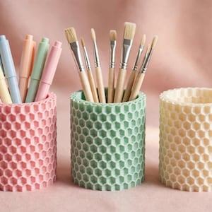 May include: Three cylindrical pencil holders with a textured honeycomb pattern. The left holder is light pink and contains pastel-coloured pens. The centre holder is mint green and holds various paintbrushes. The right holder is cream-coloured.