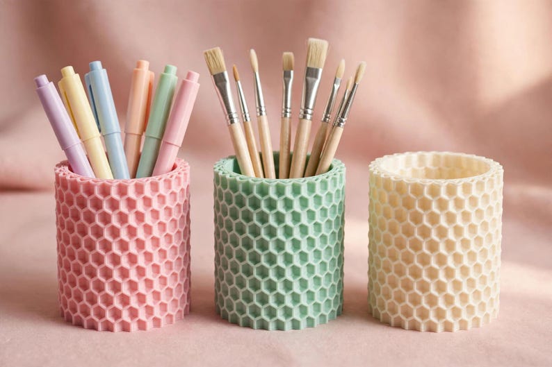 May include: Three cylindrical pencil holders with a textured honeycomb pattern. The left holder is light pink and contains pastel-coloured pens. The centre holder is mint green and holds various paintbrushes. The right holder is cream-coloured.