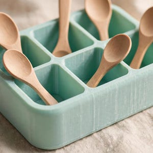 May include: A light mint green rectangular tray with six individual square compartments. Five small, light-coloured wooden spoons are placed inside the compartments, with their handles extending upwards from the tray's structure.