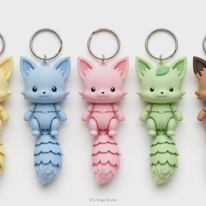 May include: Five fox-shaped keychains with metal rings, each featuring a different pastel color: yellow, light blue, pink, light green, and brown. Each figure has large eyes, pointed ears, and a segmented tail. The text STLForge Studio is at the bottom.