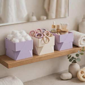 May include: Three square storage containers in light purple and cream colors. The left container holds white cotton balls, the middle container holds various hair ties, and the right container holds a glass dropper bottle, a small jar, and cotton swabs.