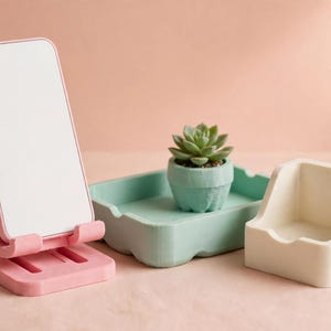 May include: A pink adjustable smartphone stand holding a white rectangular phone, a light green rectangular tray containing a small green succulent in a matching pot, and a cream-coloured square desk organiser with an open front design.