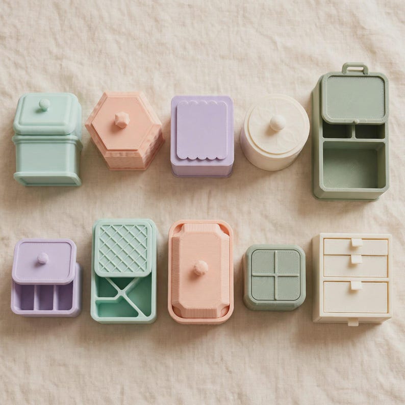 May include: A collection of ten small silicone storage containers and organizers in pastel shades of mint green, peach, lavender, and cream. The set includes various shapes like rectangular, hexagonal, and circular boxes, some with lids and compartments.