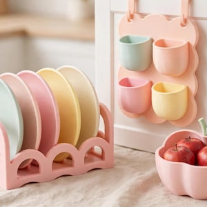 May include: A pink scalloped dish rack holding four pastel plates in mint, pink, and yellow. Beside it, a wall-mounted organiser with four pastel cups and a pink apple-shaped bowl containing three red apples, all in a soft colour palette.
