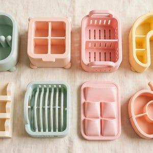 May include: A collection of eight pastel-coloured silicone organisers in various shapes and configurations. The set includes rectangular bins with dividers, a tiered shelf, a dish rack, a pocketed tray, and an apple-shaped container in shades of mint, peach, and yellow.