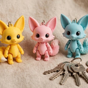 May include: Three articulated fox figurines in yellow, pink, and light blue, each featuring large dark eyes, pointed ears, and a textured tail. A metal keyring is attached to the top of each fox head, and a set of metal keys and coins sit nearby.