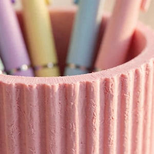 May include: A cylindrical pink pen holder with a vertical ribbed texture on the exterior. The container is filled with several pastel-coloured pens in shades of light green, purple, yellow, blue, and pink, each featuring a metallic silver band.