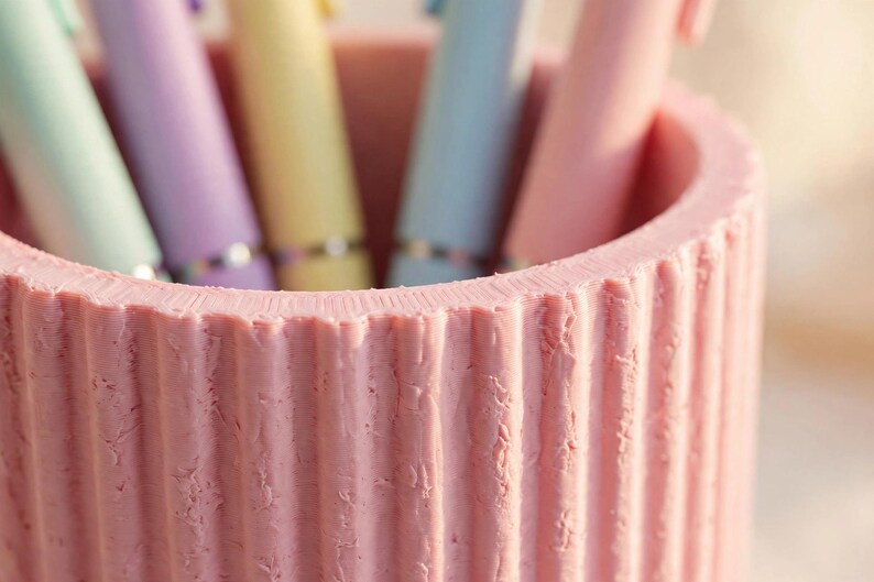 May include: A cylindrical pink pen holder with a vertical ribbed texture on the exterior. The container is filled with several pastel-coloured pens in shades of light green, purple, yellow, blue, and pink, each featuring a metallic silver band.