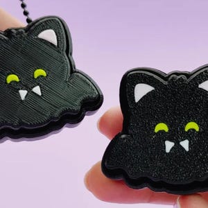 May include: Two black bat-shaped charms with white inner ears, bright yellow eyes, and two white fangs. One charm features a black ball chain attached to the top, while the other is held independently, both displaying a textured surface finish.