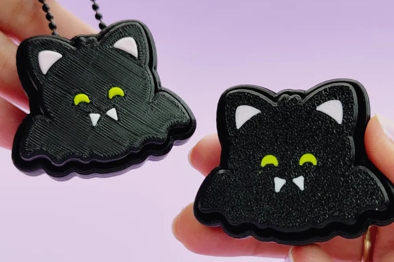 May include: Two black bat-shaped charms with white inner ears, bright yellow eyes, and two white fangs. One charm features a black ball chain attached to the top, while the other is held independently, both displaying a textured surface finish.