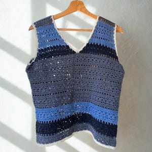 May include: A handmade crochet vest in shades of blue and gray, hanging on a wooden hanger. The sleeveless top features a V-neck and horizontal stripes of varying blue tones. The edges are finished with a cream-colored trim.