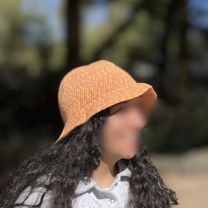 Op de afbeelding: Een gebreide vissershoed in tinten oranje en geel. De hoed heeft een licht gebogen rand en een getextureerd uiterlijk. De hoed is gemaakt van een zacht, lichtgewicht materiaal, perfect voor bescherming tegen de zon. Een stijlvol accessoire.