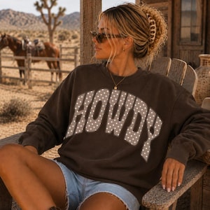 May include: A dark brown long-sleeve sweatshirt featuring the word HOWDY in large, arched, light-colored letters with a white polka dot pattern across the chest. The garment has a crew neckline and a relaxed fit, constructed from a solid fabric.
