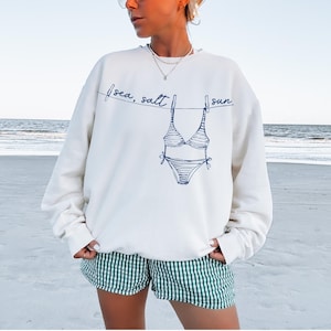 May include: Cream-colored sweatshirt with a blue line drawing of a bikini hanging on a clothesline. The words "sea, salt, sun" are written above the bikini. The person is wearing green and white checkered shorts.