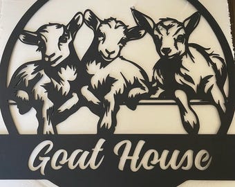 May include: Black metal sign with a circular cutout featuring three goats peeking over a fence. The sign reads "Goat House" in a stylized font.