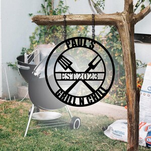 May include: Black metal sign with the text "Paul's Grill N Chill Est 2023" and crossed grill tools. The sign is hanging from a chain.