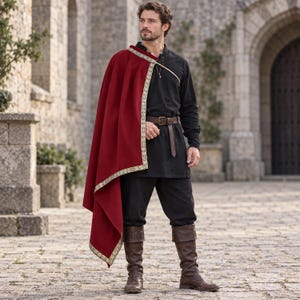 Könnte beinhalten: Ein dunkelroter Umhang mit goldener Musterborte über einer Schulter, getragen über einer schwarzen langärmeligen Tunika und einer schwarzen Hose. Ein brauner Ledergürtel mit Metallschnalle ist in der Taille befestigt, dazu braune Lederstiefel.