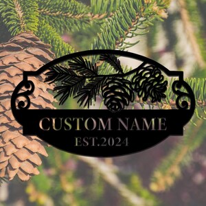 Könnte beinhalten: Schwarzes Metallschild mit einem Design aus Kiefernzweig und Tannenzapfen. Das Schild trägt die Aufschrift "CUSTOM NAME EST. 2024".