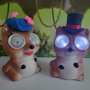 May include: Two ceramic animal figurines with light-up eyes. The figurine on the left is wearing a blue hat with a pink flower. The figurine on the right is wearing a black top hat with a red band.