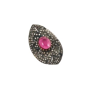 May include: An oval-shaped silver ring with a large, oval, pink gemstone at the center. The ring is encrusted with small, clear gemstones and features an ornate, filigree design around the pink stone.