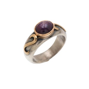 May include: A silver ring with a purple gemstone set in a gold bezel. The ring has a textured band with a gold design.