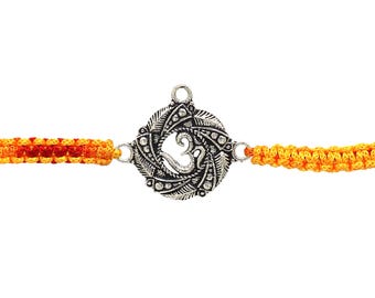 May include: A silver-colored bracelet featuring an Om symbol in the center, surrounded by a decorative design. The bracelet has orange and yellow woven cords on each side, creating a vibrant contrast against the silver.
