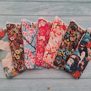 Puede incluir: Una colección de siete estuches rectangulares de tela con diversos patrones florales, botánicos y tradicionales. Cada estuche se asegura con un pequeño botón en el borde superior, mostrando diseños que incluyen flores de cerezo, flores oscuras y una geisha.
