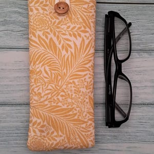 May include: A yellow floral print glasses case with a wooden button closure. Next to the case are black framed eyeglasses. The case is made of fabric and has a cream-colored top.