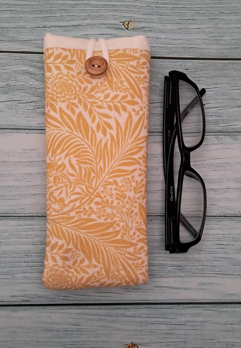 May include: A yellow floral print glasses case with a wooden button closure. Next to the case are black framed eyeglasses. The case is made of fabric and has a cream-colored top.
