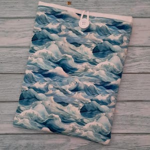 May include: A rectangular fabric sleeve featuring a repeating pattern of blue and white ocean waves. The sleeve has a white top edge and is secured with a white elastic loop that fastens over a single white button centred on the upper portion.