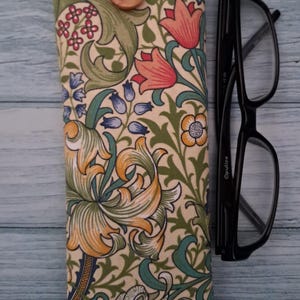 May include: A floral patterned glasses case with a red trim and wooden button closure. The case features a cream background with orange, green, and blue floral designs. Black eyeglasses rest beside the case.