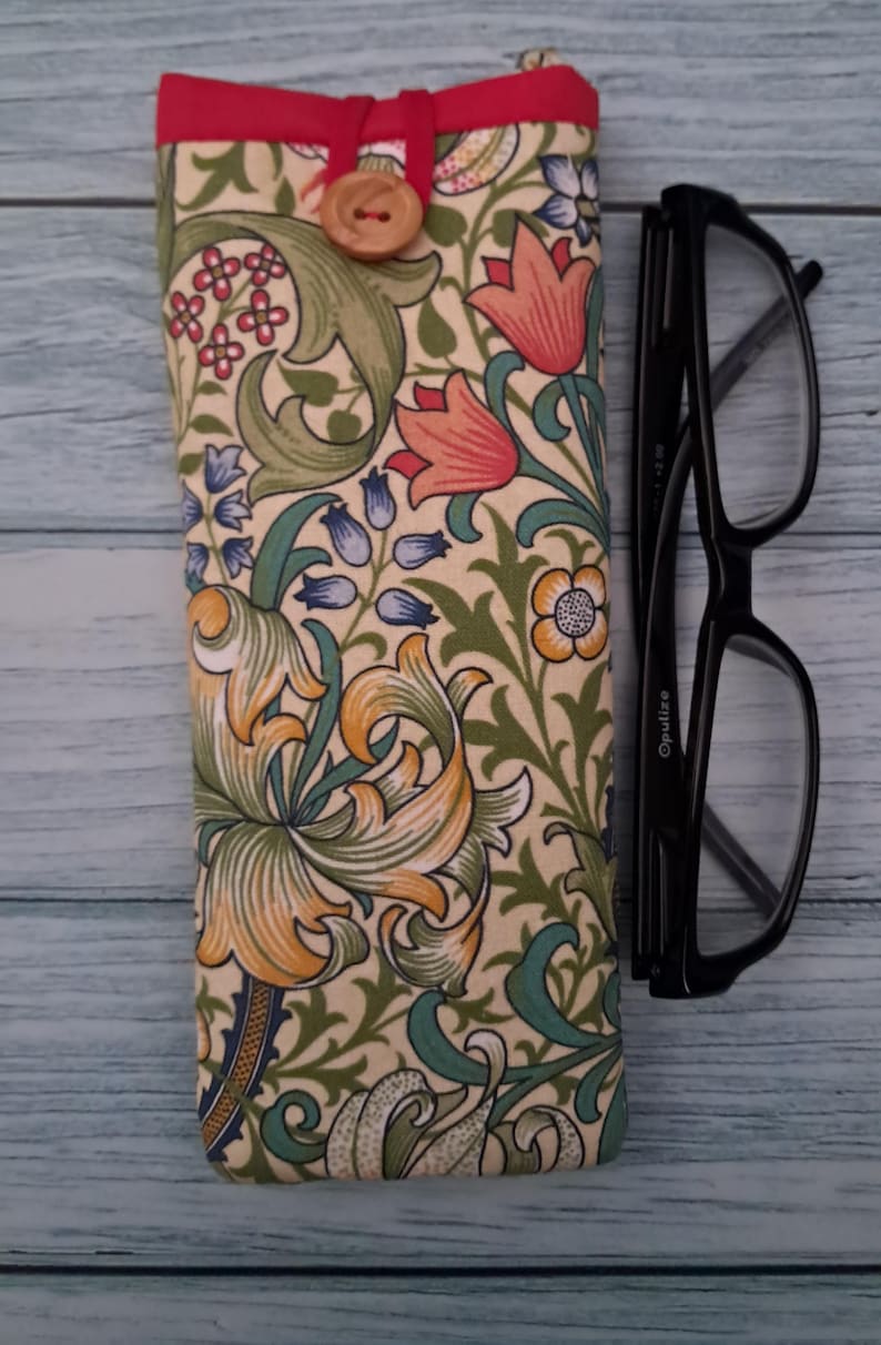 May include: A floral patterned glasses case with a red trim and wooden button closure. The case features a cream background with orange, green, and blue floral designs. Black eyeglasses rest beside the case.