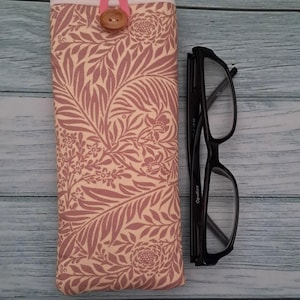 May include: A fabric glasses case with a floral pattern in shades of pink and cream, secured with a pink loop and a wooden button. Next to it are black-framed eyeglasses.