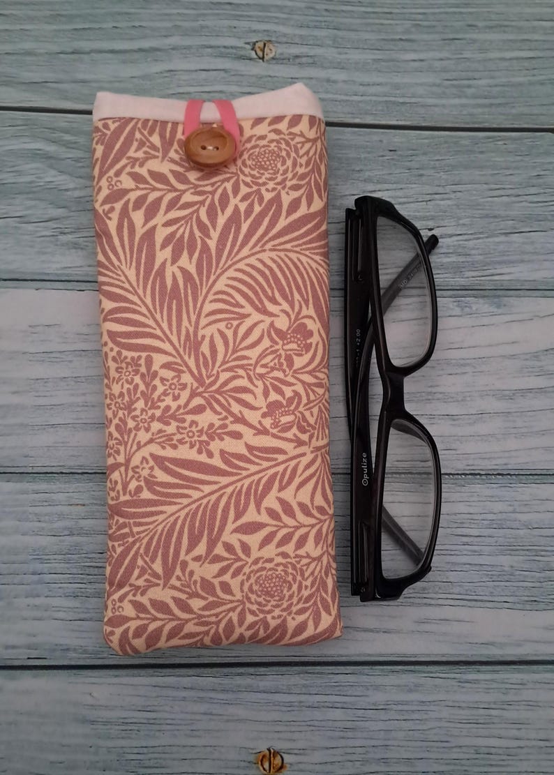 May include: A fabric glasses case with a floral pattern in shades of pink and cream, secured with a pink loop and a wooden button. Next to it are black-framed eyeglasses.