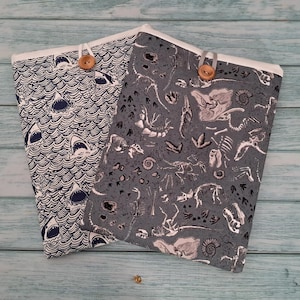 Puede incluir: Dos fundas rectangulares de tela con cierre de botón. Una funda presenta un patrón azul oscuro y blanco de tiburones en olas. La otra funda es gris con un estampado blanco de esqueletos de dinosaurios, fósiles y huellas en la superficie.