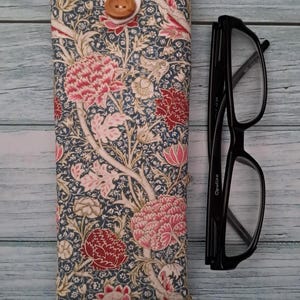 May include: A fabric glasses case with a floral pattern in shades of pink, red, and cream on a navy blue background. The case is closed with a wooden button and loop. Black eyeglasses are next to the case.