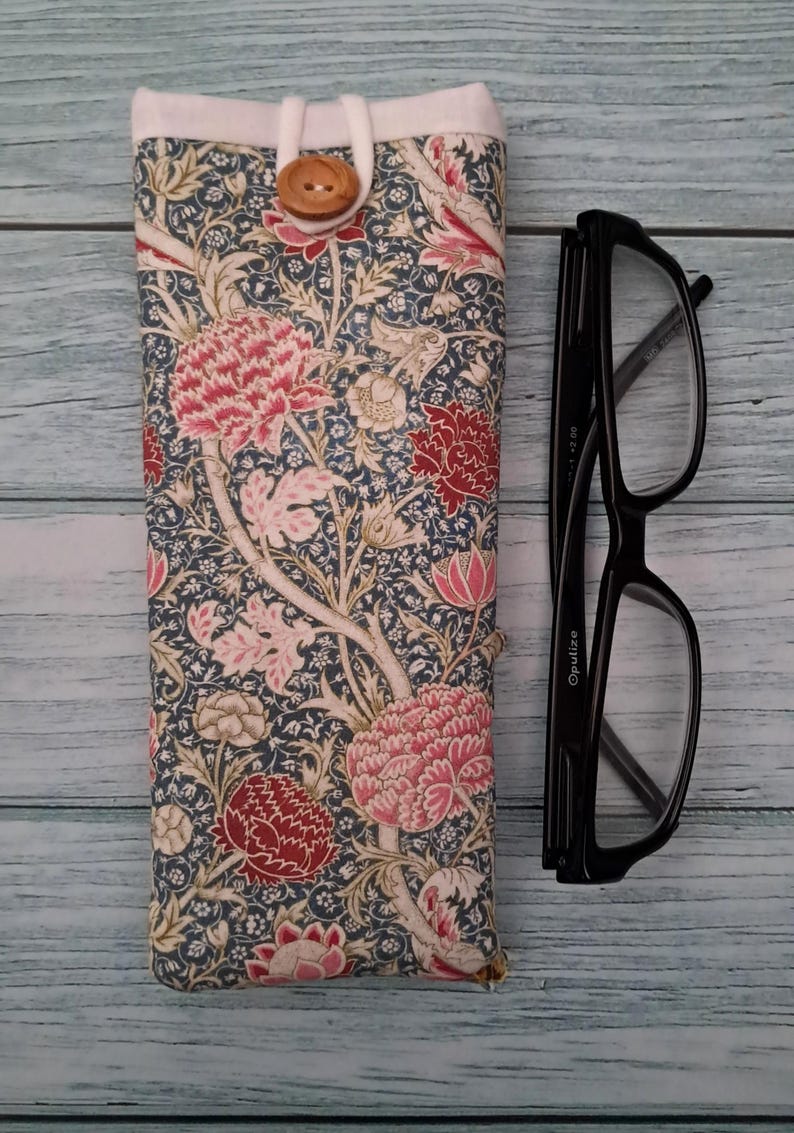 May include: A fabric glasses case with a floral pattern in shades of pink, red, and cream on a navy blue background. The case is closed with a wooden button and loop. Black eyeglasses are next to the case.