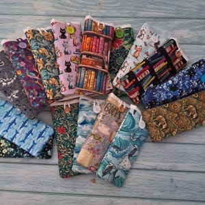 Puede incluir: Una colecci&oacute;n de estuches rectangulares de tela con varios patrones, incluyendo gatos, estanter&iacute;as, motivos florales, ballenas y dise&ntilde;os abstractos. Cada estuche se asegura con un peque&ntilde;o bot&oacute;n y est&aacute; dispuesto en forma de abanico.