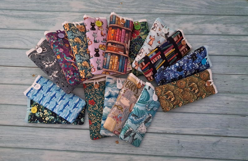 Puede incluir: Una colecci&oacute;n de estuches rectangulares de tela con varios patrones, incluyendo gatos, estanter&iacute;as, motivos florales, ballenas y dise&ntilde;os abstractos. Cada estuche se asegura con un peque&ntilde;o bot&oacute;n y est&aacute; dispuesto en forma de abanico.