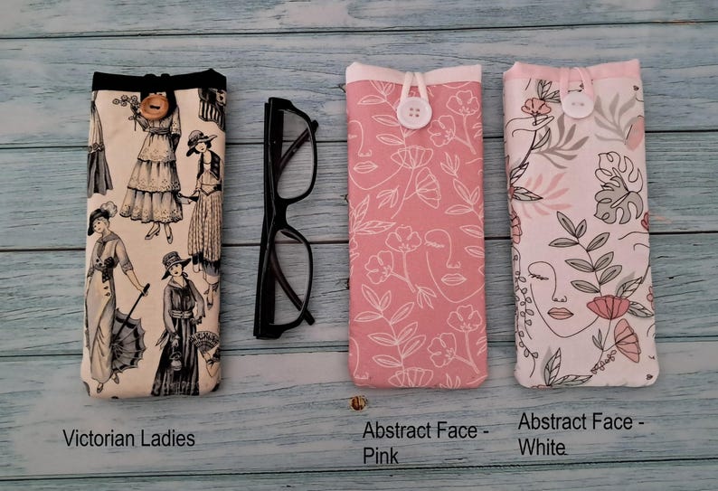 Puede incluir: Tres estuches rectangulares de tela para gafas con cierre de bot&oacute;n y un par de gafas negras. El primer estuche muestra Victorian Ladies, el segundo es Abstract Face - Pink y el tercero es Abstract Face - White con dibujos lineales florales.