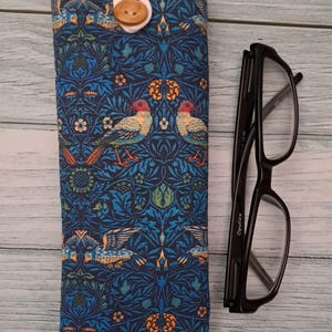 May include: A blue fabric glasses case with a bird and floral pattern, a white top, and a wooden button closure. Black eyeglasses are next to the case on a light wood surface.
