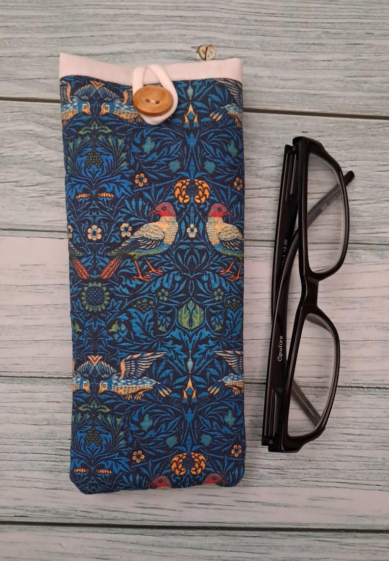 May include: A blue fabric glasses case with a bird and floral pattern, a white top, and a wooden button closure. Black eyeglasses are next to the case on a light wood surface.