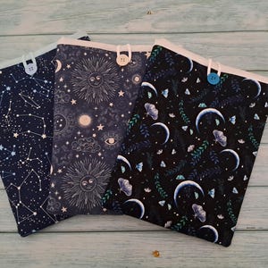 Puede incluir: Tres fundas de libros de tela con diseños celestiales. Una presenta constelaciones, otra un sol y estrellas, y la tercera lunas y mariposas. Cada funda tiene una parte superior blanca y un cierre de botón.
